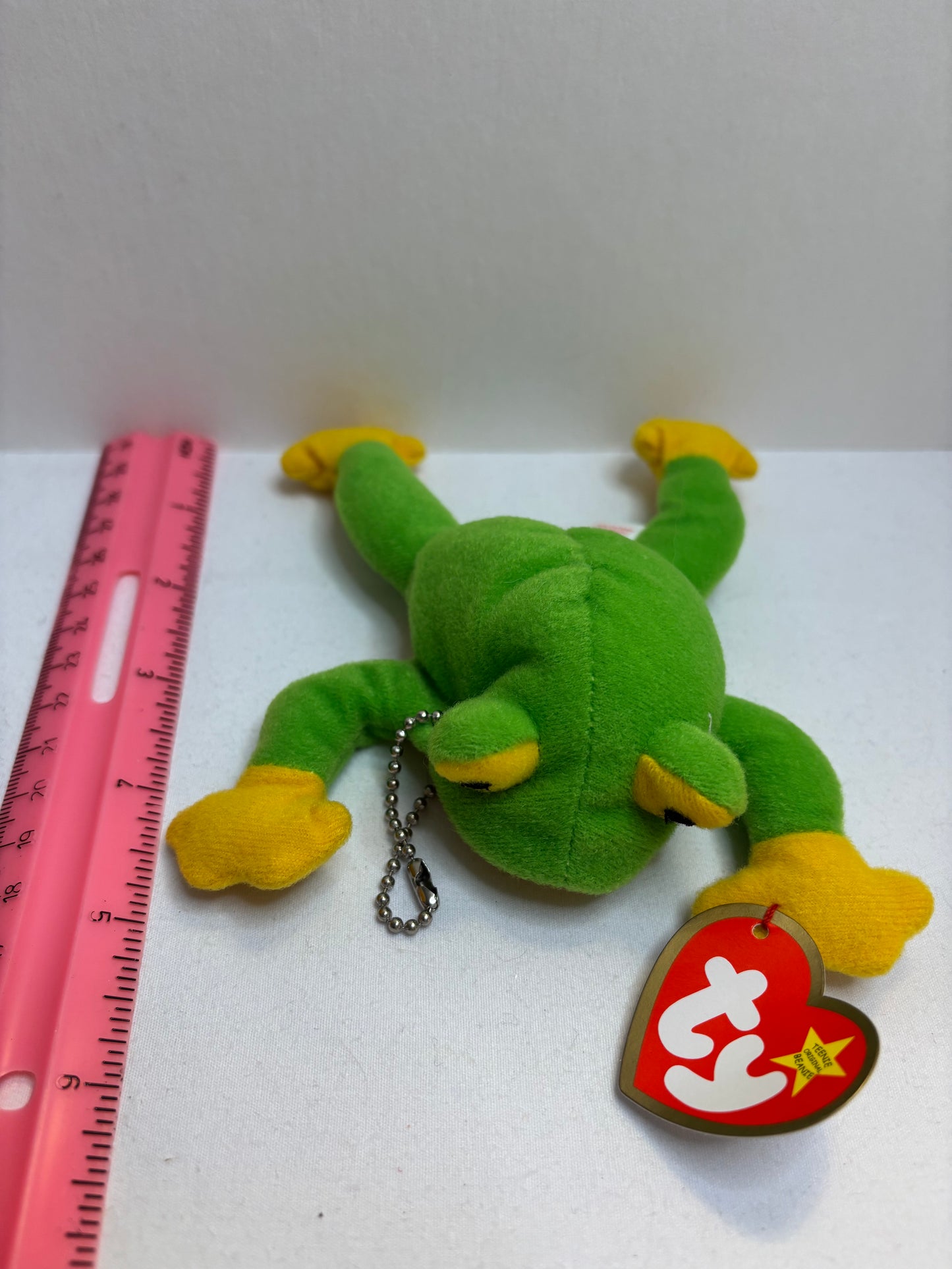 Green plush frog bag charm with yellow feet and a red tag on a white background